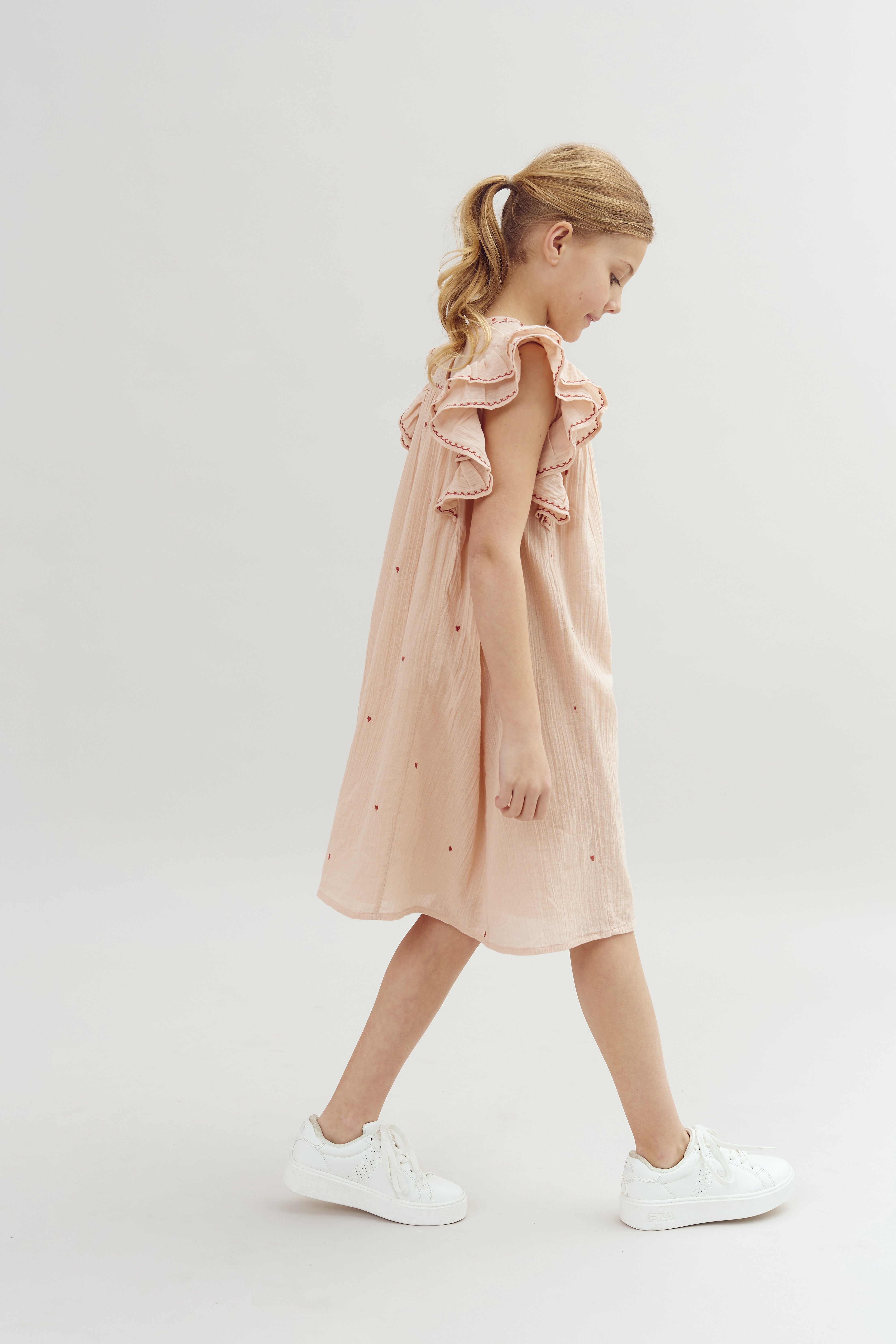 Mädchen im rosa Kleid mit Rüschen läuft, helle Haare zu einem Pferdeschwanz gebunden, weiße Turnschuhe tragend, auf hellem, neutralem Hintergrund.