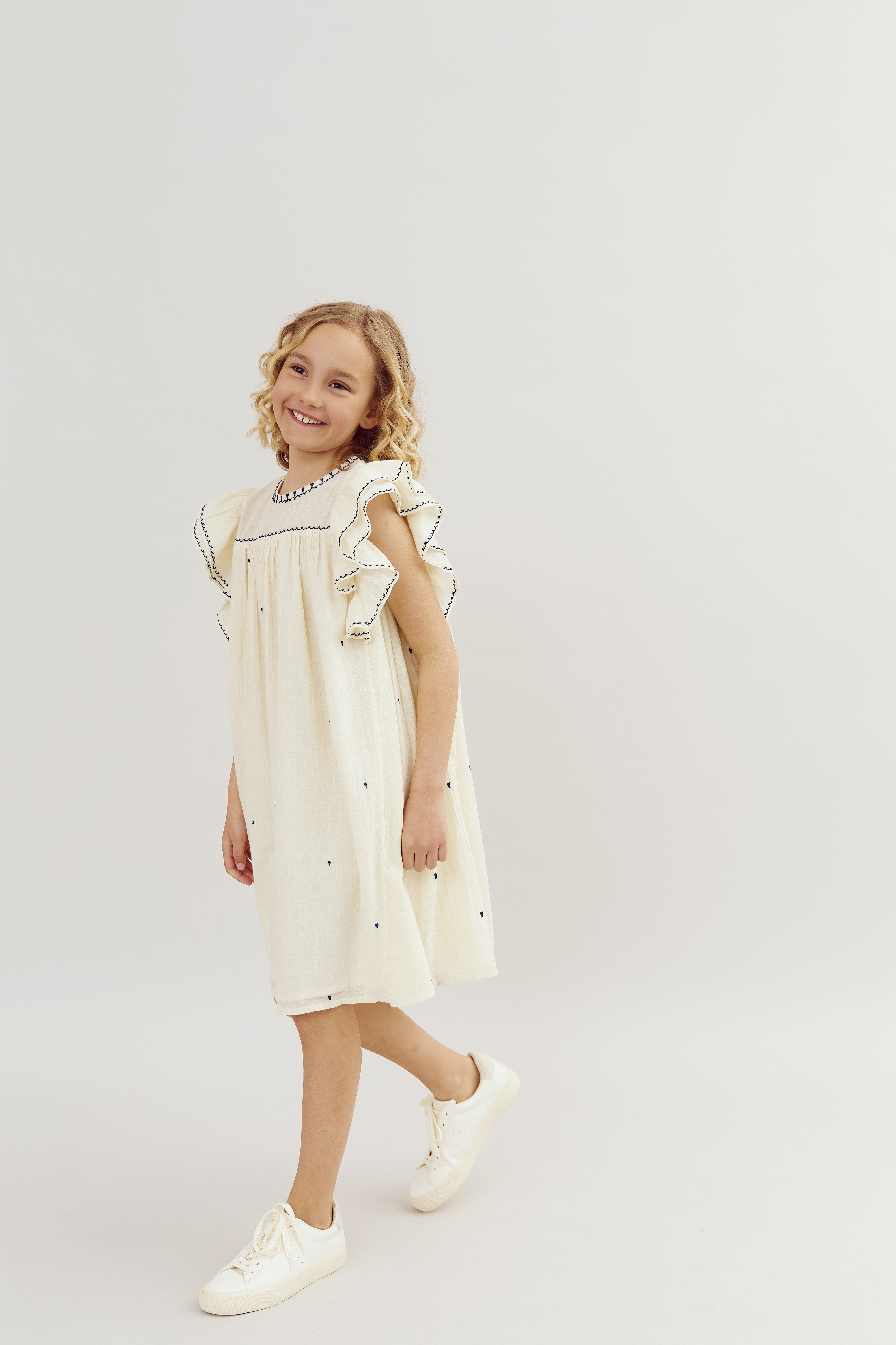 Ein Mädchen in einem cremefarbenen Kleid mit Rüschen geht lächelnd nach rechts. Sie trägt weiße Turnschuhe. Der Hintergrund ist einfarbig hell und leer.